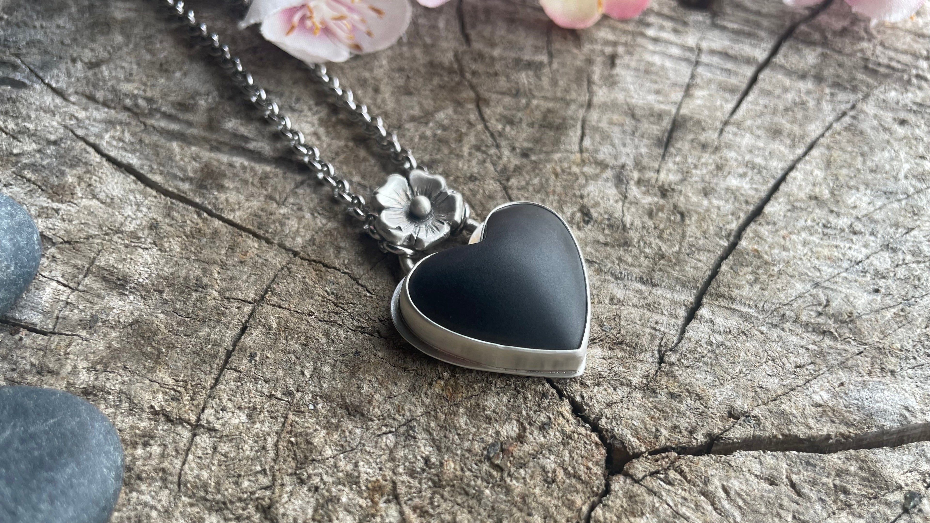 Heart-shaped black pendant on a silver chain on a wooden surface with flowers.